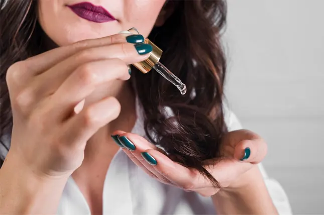 A woman with dark hair and bold lipstick drips serum from a dropper onto her hand, preparing to apply it to her hair.