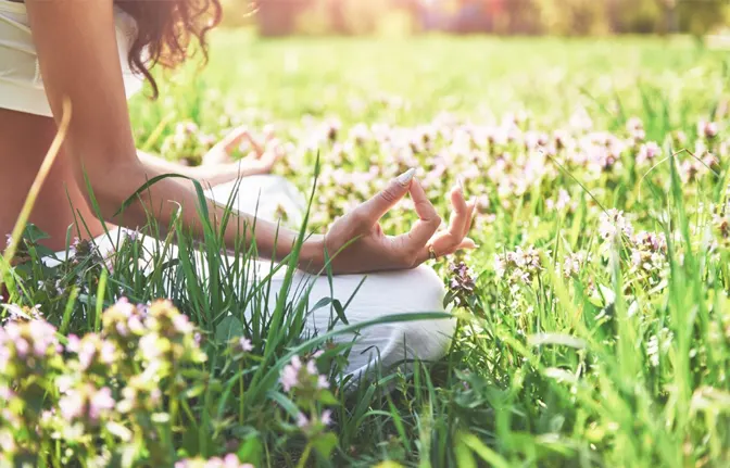 A person meditating in a sunny field, surrounded by green grass and blooming flowers, with hands resting in a serene pose.