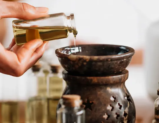 A hand pours essential oil from a glass vial into a dark ceramic oil diffuser, surrounded by various glass bottles of oils
