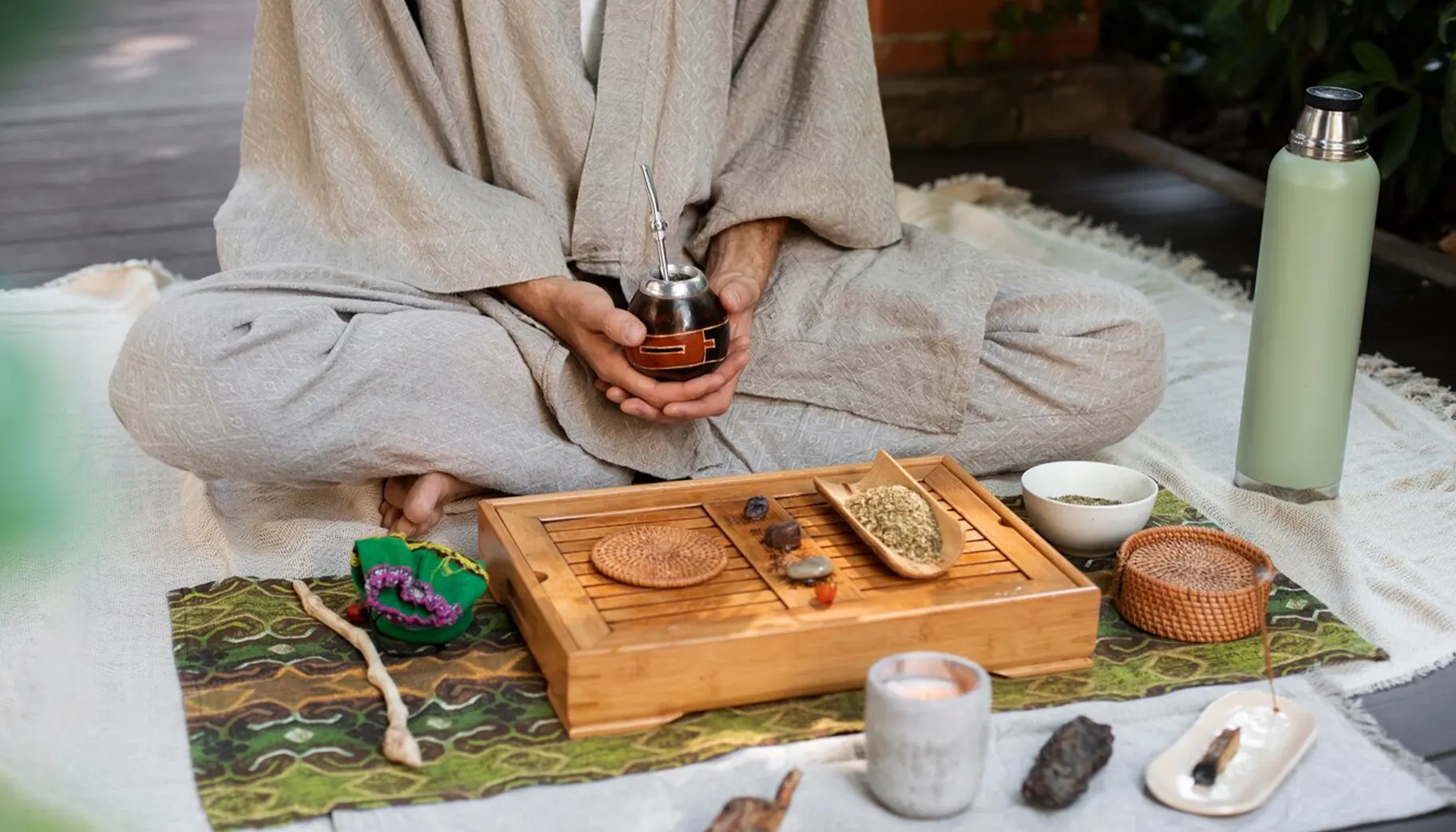 A person in a gray robe sits cross-legged, holding a mate cup, surrounded by herbal items and a candle on a patterned cloth.