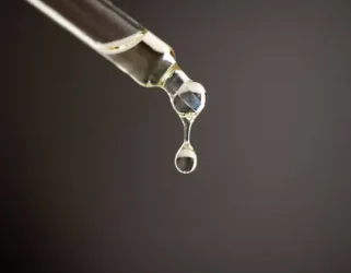 Close-up of a glass dropper releasing a clear droplet against a dark background, showcasing the fluid's texture and clarity